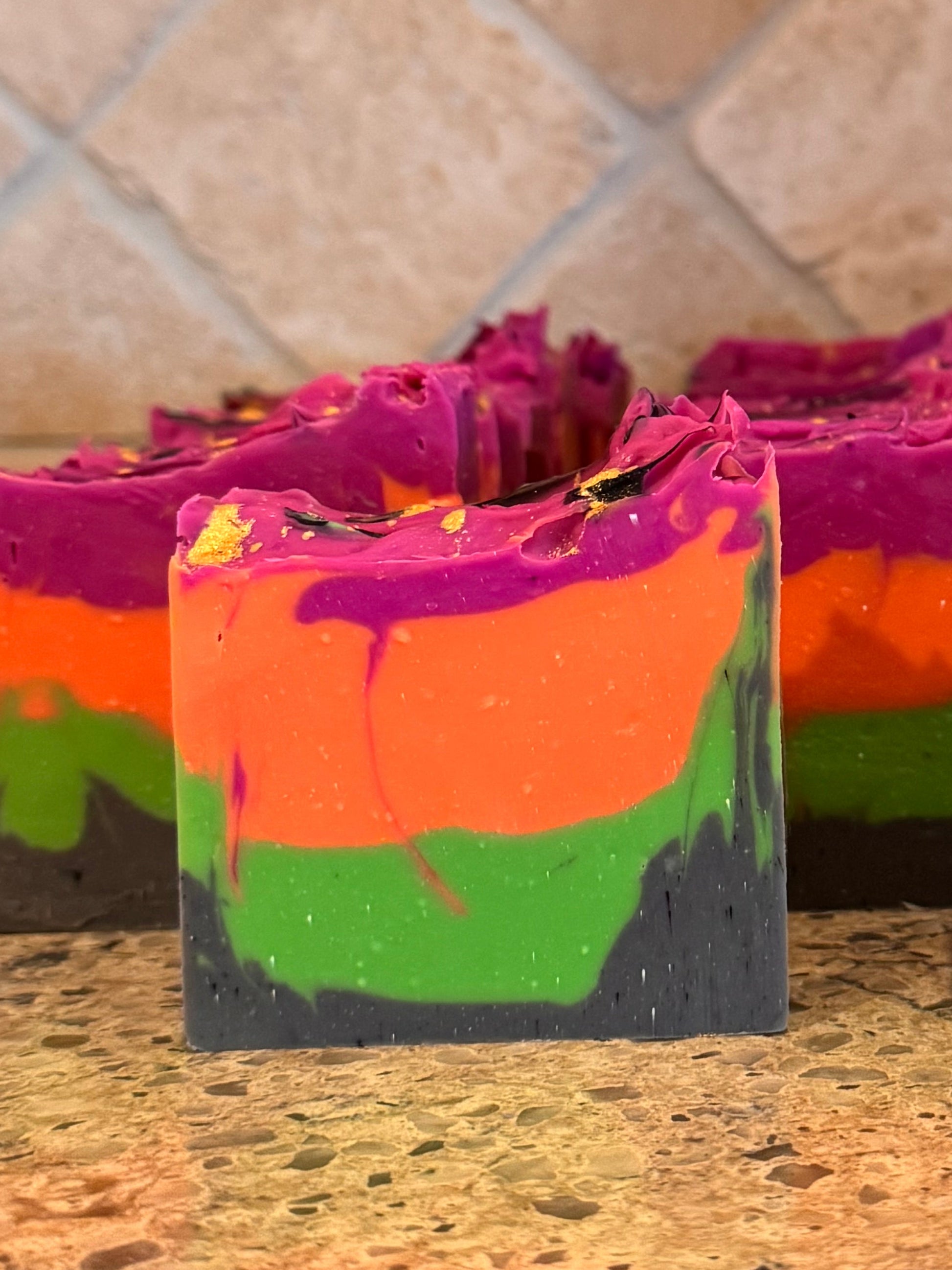 Five rectangular layered soap bars with a rainbow color scheme of purple, orange, green, and dark gray, arranged on a speckled granite countertop with a tiled backsplash behind them.